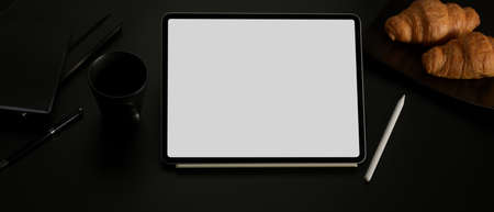 Cropped shot of blank screen tablet on dark office desk with coffee, croissants and stationeryの写真素材