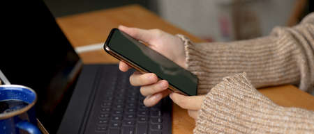 Side view of female hands holding smartphone while sitting at workspace with digital tablet on wooden tableの写真素材
