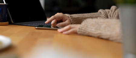 Side view of female touching on smartphone while sitting at workspace with digital tablet and copy...