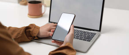 Cropped shot of female hands holding mock up smartphone while sitting at worktable with mock up...