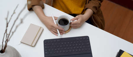 Overhead shot of female holding coffee cup on white office desk with office supplies