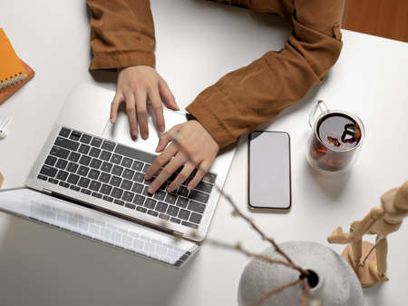 Overhead shot of female typing on laptop keyboard on white table with mock up smartphone, supplies...