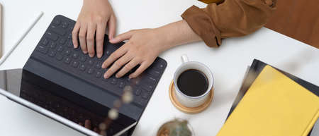 Overhead shot of female typing on tablet keyboard on worktable with coffee cup, books and cactus pot