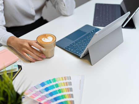 Cropped shot of female sitting at workspace, holding a glass of coffee while concentrating on her...