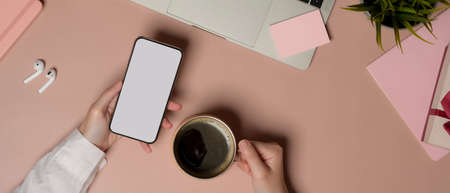 Overhead shot of female using smartphone and holding coffee cup on pink table