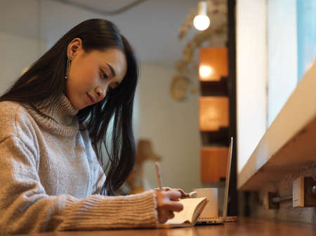 Close up view of female taking note while working with mock up laptop on wooden counter bar in cafe