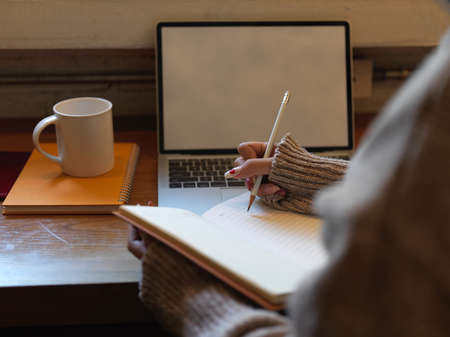 Close up view of female writing no blank notebook while siting at wooden worktable with mock up...