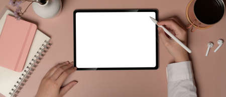 Overhead shot of female working with digital tablet on pink office desk with notebooks and...