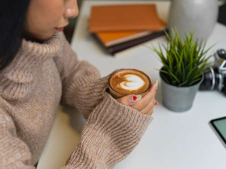 Side view of female in sweater holding a cup of hot latte coffee on office desk in office roomの写真素材