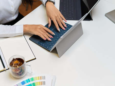 Cropped shot of female hands typing on tablet keyboard on white tablet with notebook, coffee cup and...