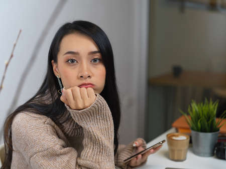 Portrait of female in sweater sitting in glass partition office room and using smartphone