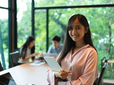 Portrait of female office worker holding digital tablet and stylus pen while standing in meeting roomの写真素材