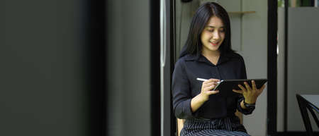 Portrait of female offie worker using digital tablet with stylus while standing against glass wall...
