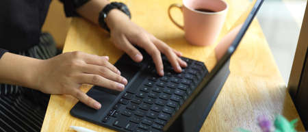 Cropped shot of female hands typing on tablet keyboard on wooden table with coffee mug in cafe