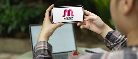 Close up view of female holding horizontal mock up smartphone while working with mock up laptop in...