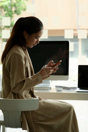Portrait of female entrepreneur using smartphone while at office desk with computer device in office...