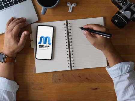 Overhead shot of male office worker using mock up smartphone while taking note on blank notebook on wooden tableの写真素材