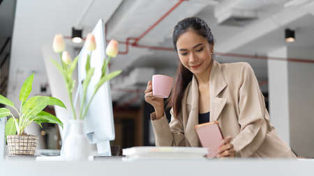 Portrait of female office worker relaxing with smartphone and a cup of coffee while working in office roomの写真素材