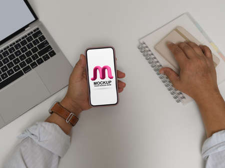 Overhead shot of male office worker holding mock up smartphone while working with laptop and schedule booksの写真素材
