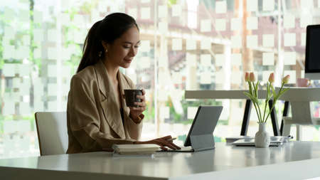 Side view of female entrepreneur working with digital tablet and holding coffee cup in office roomの写真素材
