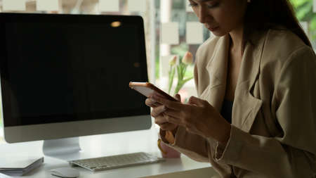 Cropped shot of female entrepreneur using smartphone while at office desk with computer device in office roomの写真素材