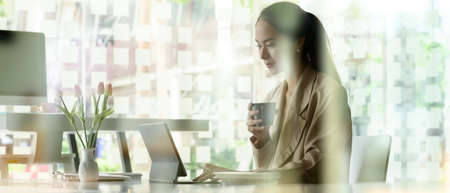Side view of female office worker holding coffee cup while working with digital tablet on white...