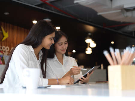 Side view of two female university students doing homework together with digital tablet and stationery in libraryの写真素材