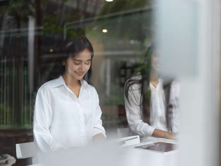 View through glass window of female concentrating on digital tablet while sitting beside her friend...