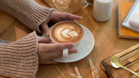 Cropped shot of young female holding latte art coffee on wooden table with stationeries