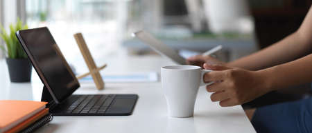 Cropped shot of young designer working with tablet while holding coffee cup in workspace