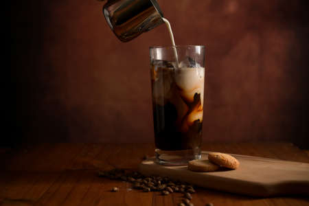 Close up view of a glass of ice coffee while adding milk with biscuit and coffee beans on wooden...