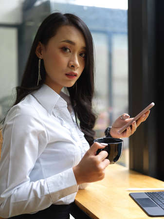 Portrait of female office worker looking into camera while relaxing with coffee cup and smartphoneの写真素材