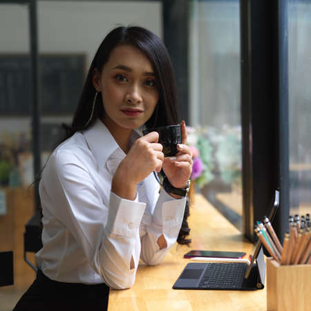 Portrait of female looking into camera while drinking coffee at portable workspace in cafeの写真素材