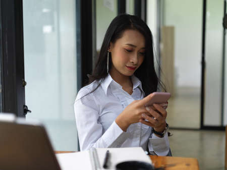 Portrait of female texting on smartphone while sitting at worktable in office roomの写真素材
