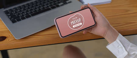 Close up view of female hand holding mock up horizontal smartphone while sitting at worktable with...