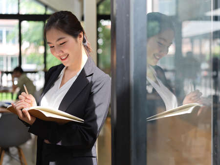 Portrait of businesswoman taking note on schedule book while standing in glass partition office roomの写真素材