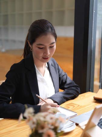Portrait of businesswoman working with laptop and taking note on blank notebook in cafeの写真素材