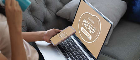 Side view of female working with mock up laptop and smartphone while sitting on couch in living room