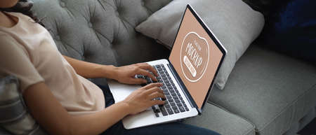 Side view of female using typing on mock up laptop keyboard on her lap while relaxed sitting on sofa