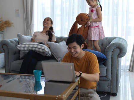 Happy family in living room, mother using digital tablet and father working with laptop while daughter playing with stuffed toy on sofaの写真素材