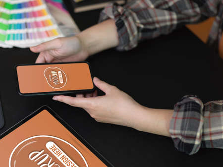 Top view of female designer using mock up smartphone on black table with mock up tablet and supplies