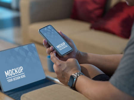Cropped shot of male hands using mock up smartphone while working with mock up tablet in living room