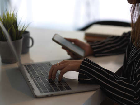 Side view of female hand typing on laptop and using smartphone on worktable in office roomの写真素材