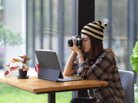 Side view of female taking photo white digital camera while sitting at coffee table with laptop in cafeの写真素材