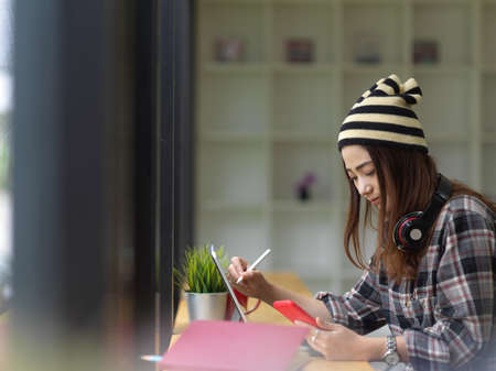 Side view of female teenager doing homework with digital tablet and supplies on wooden table in cafeの写真素材