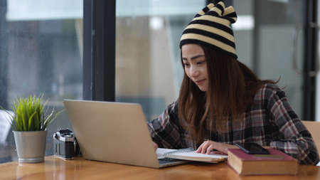 Portrait of female student doing assignment with laptop, stationery and textbook in cafe