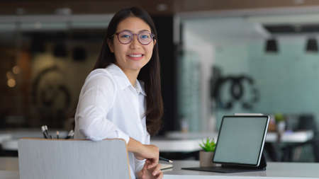 Portrait of female office worker smiling and looking into camera while working with mock up tablet in officeの写真素材