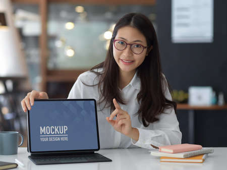 Young pretty businesswoman pointing at blank screen mock up tablet in the office room, include clippingの写真素材