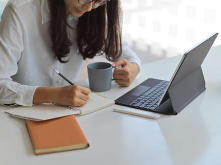 Cropped shot young businesswoman writing her project while holding a cup of coffee in office room