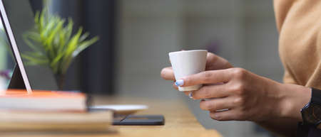 Side view of female hands holding coffee cup while working on wooden bar with tablet and book